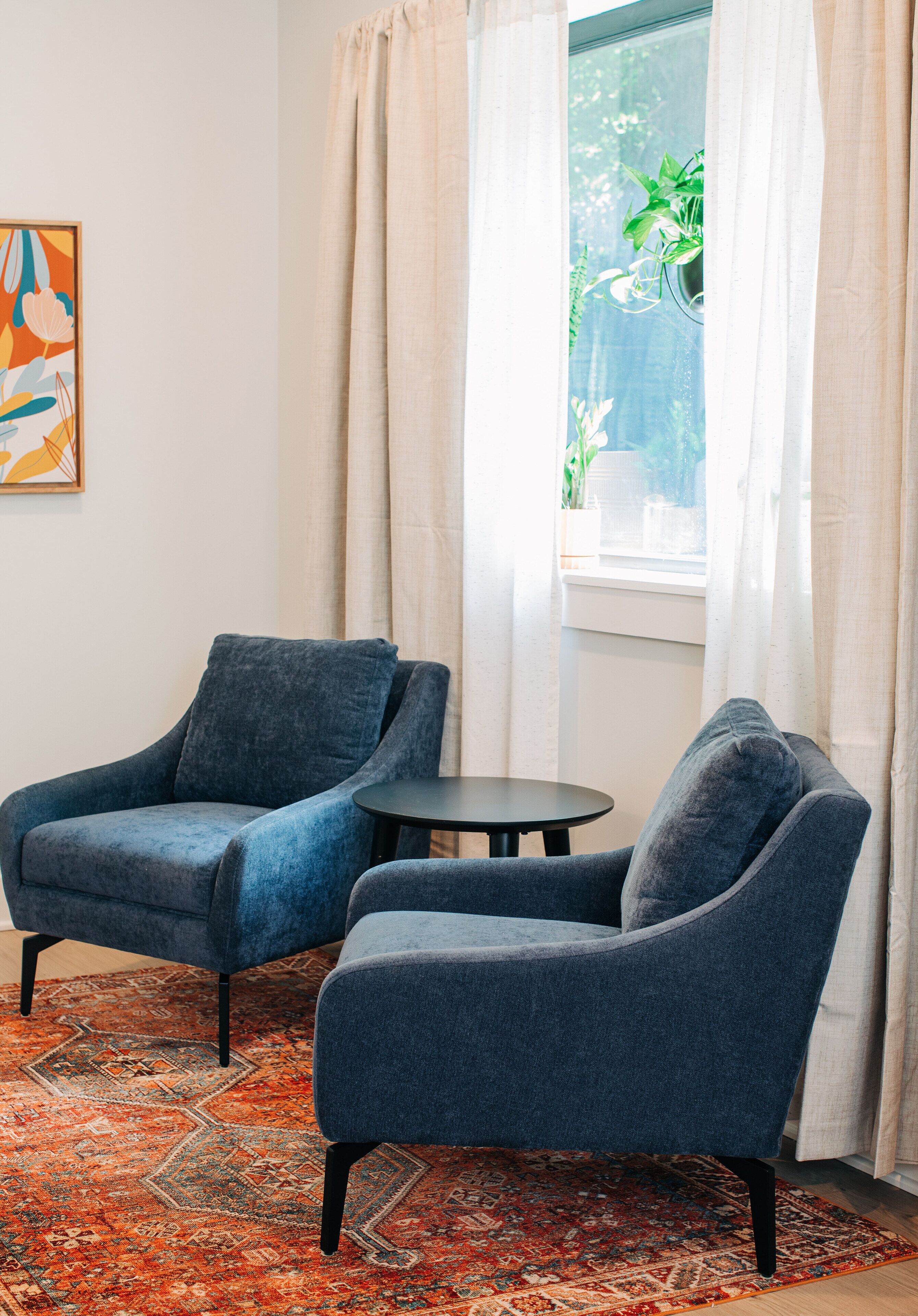 A room with two blue armchairs, a small round table, a patterned rug, and a window with curtains.