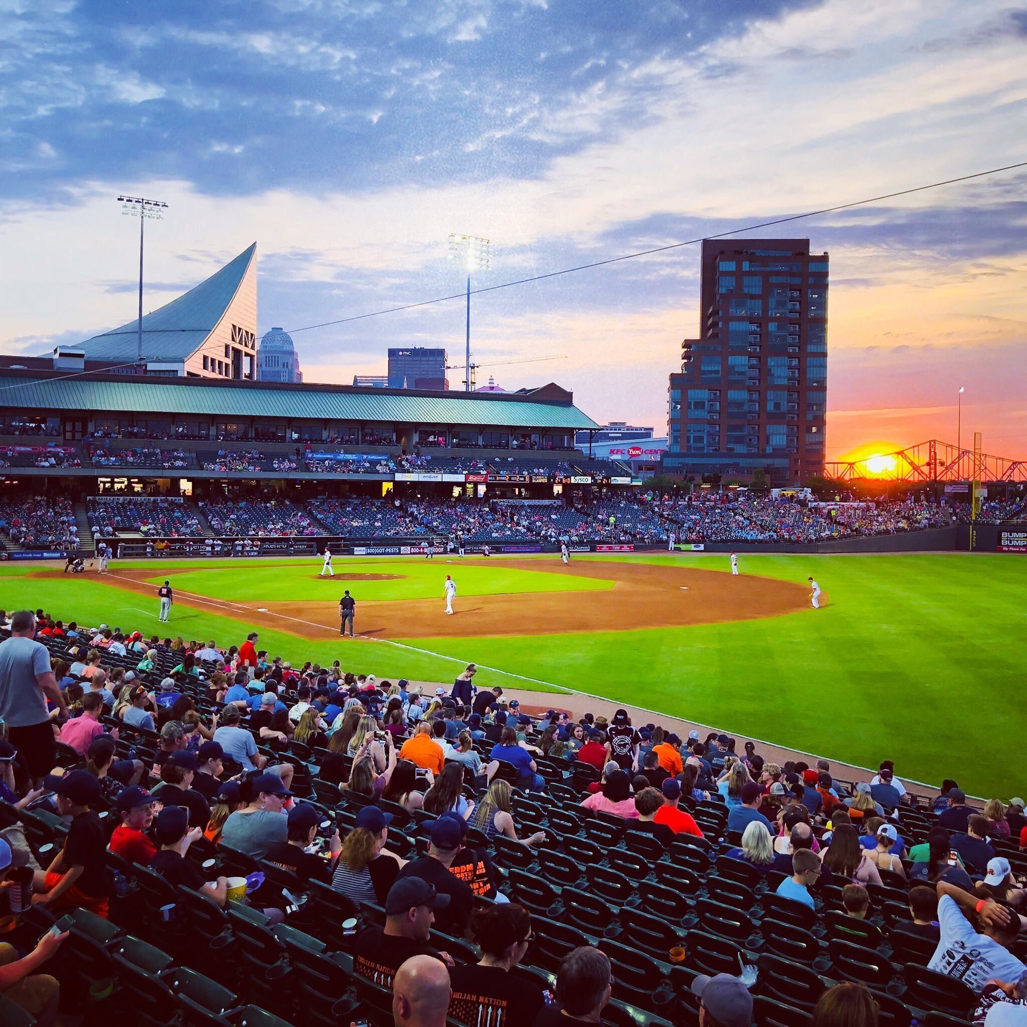 A baseball stadium with a large crowd, a clear sky, and a sunset.