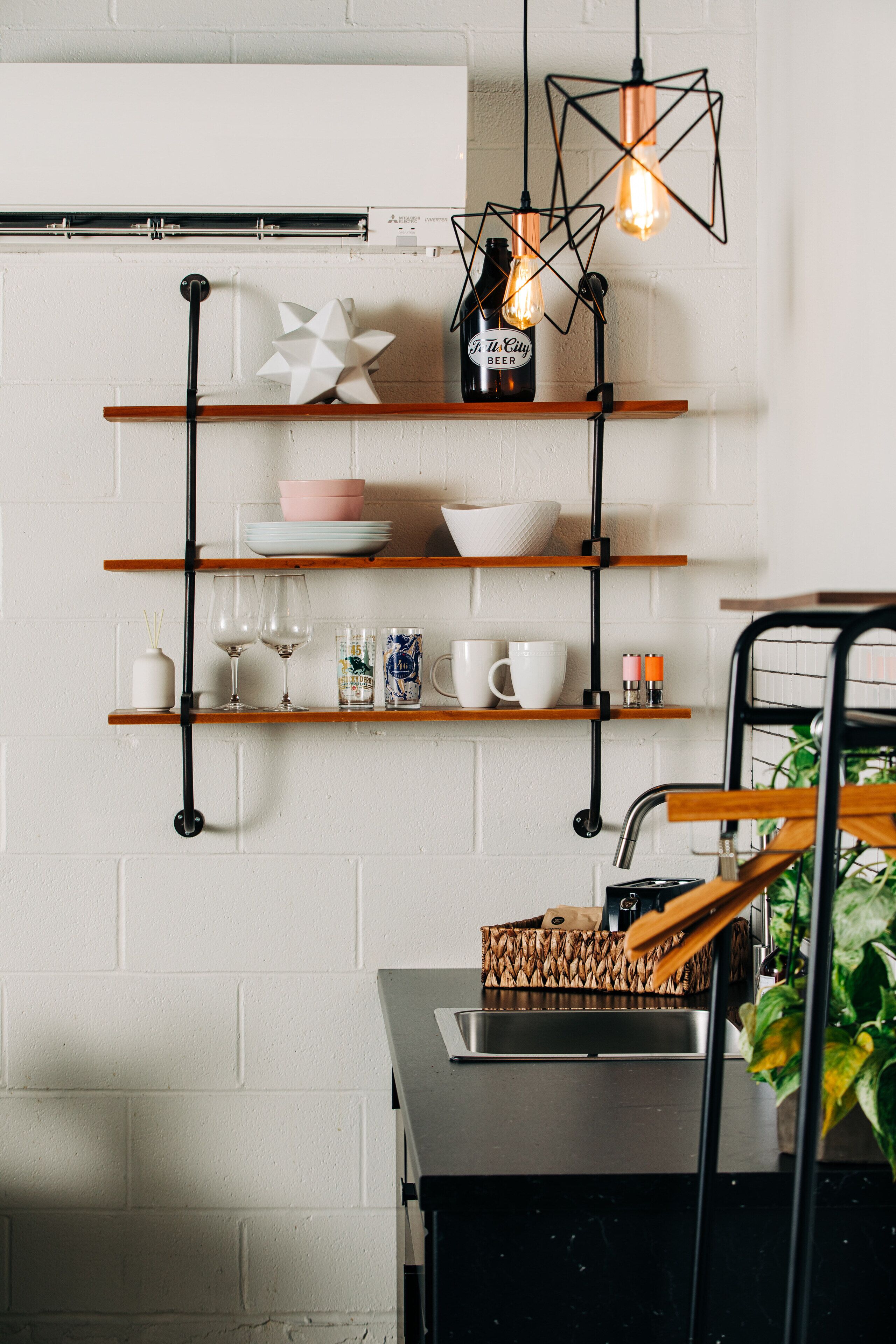 A kitchen with wooden shelves holding dishes, glasses, and a bottle of beer. There is a hanging light fixture and a plant on the counter.
