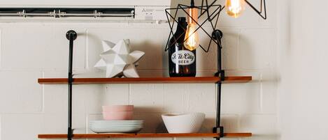 A kitchen with wooden shelves holding dishes, glasses, and a bottle of beer. There is a hanging light fixture and a plant on the counter.