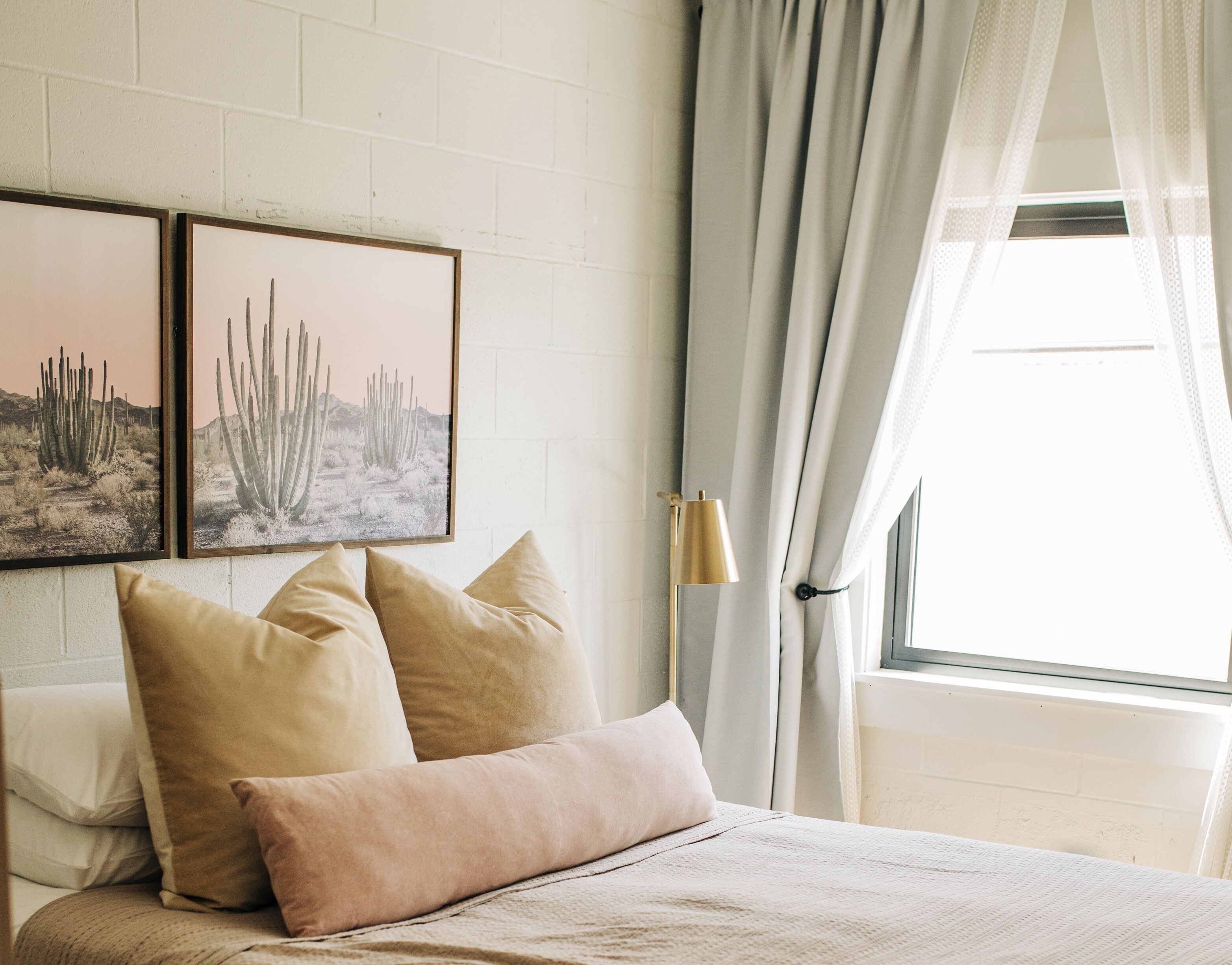 A bedroom with a bed, two framed desert landscape pictures, a bedside lamp, and a window with curtains.