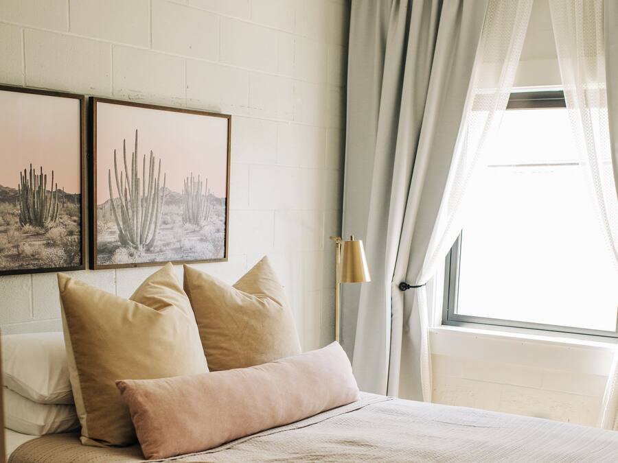 A bedroom with a bed, two framed desert landscape pictures, a bedside lamp, and a window with curtains.