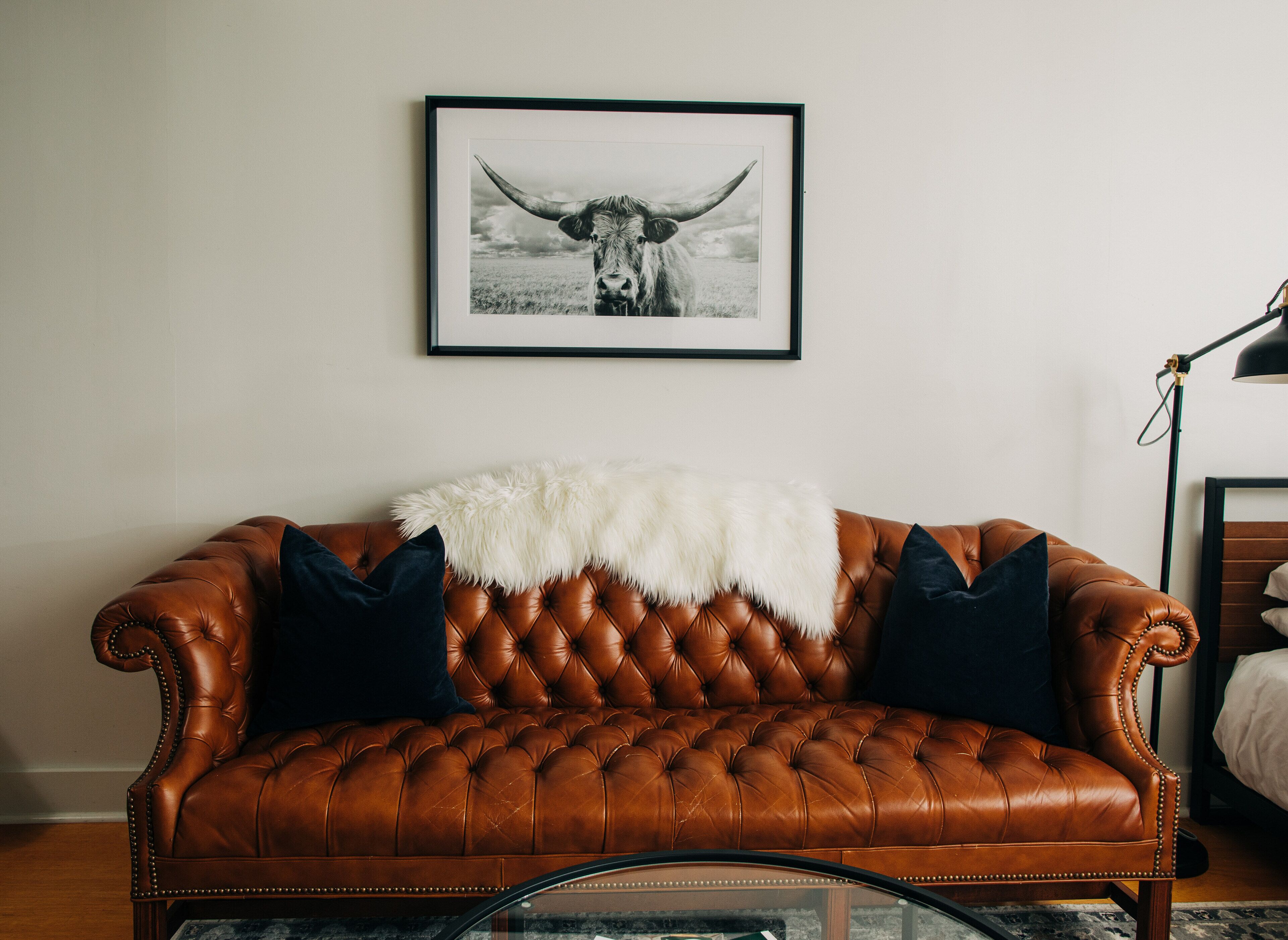 A brown leather sofa with a white furry throw blanket and two dark pillows, positioned against a white wall with a framed black and white photograph above it.