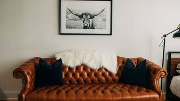 A brown leather sofa with a white furry throw blanket and two dark pillows, positioned against a white wall with a framed black and white photograph above it.