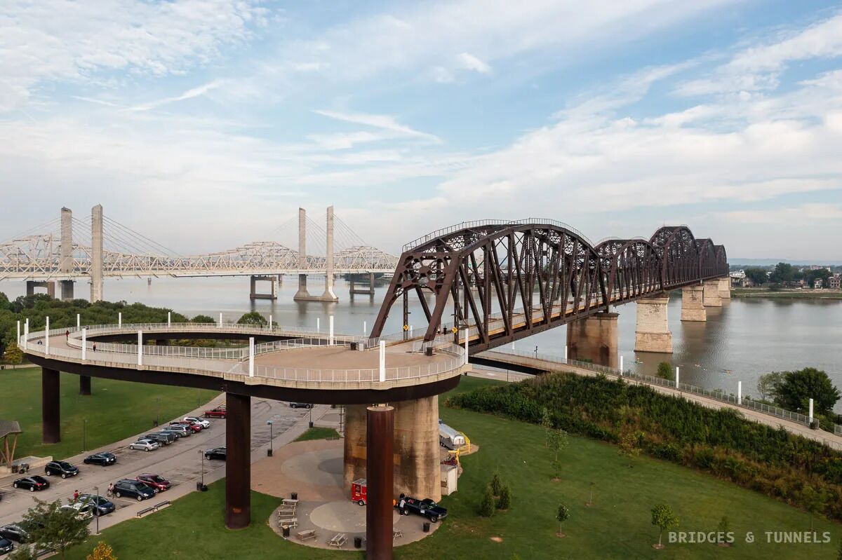 Uma grande ponte de aço com passarela para pedestres sobre um rio, cercada por vegetação e um estacionamento.