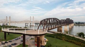 Uma grande ponte de aço com passarela para pedestres sobre um rio, cercada por vegetação e um estacionamento.
