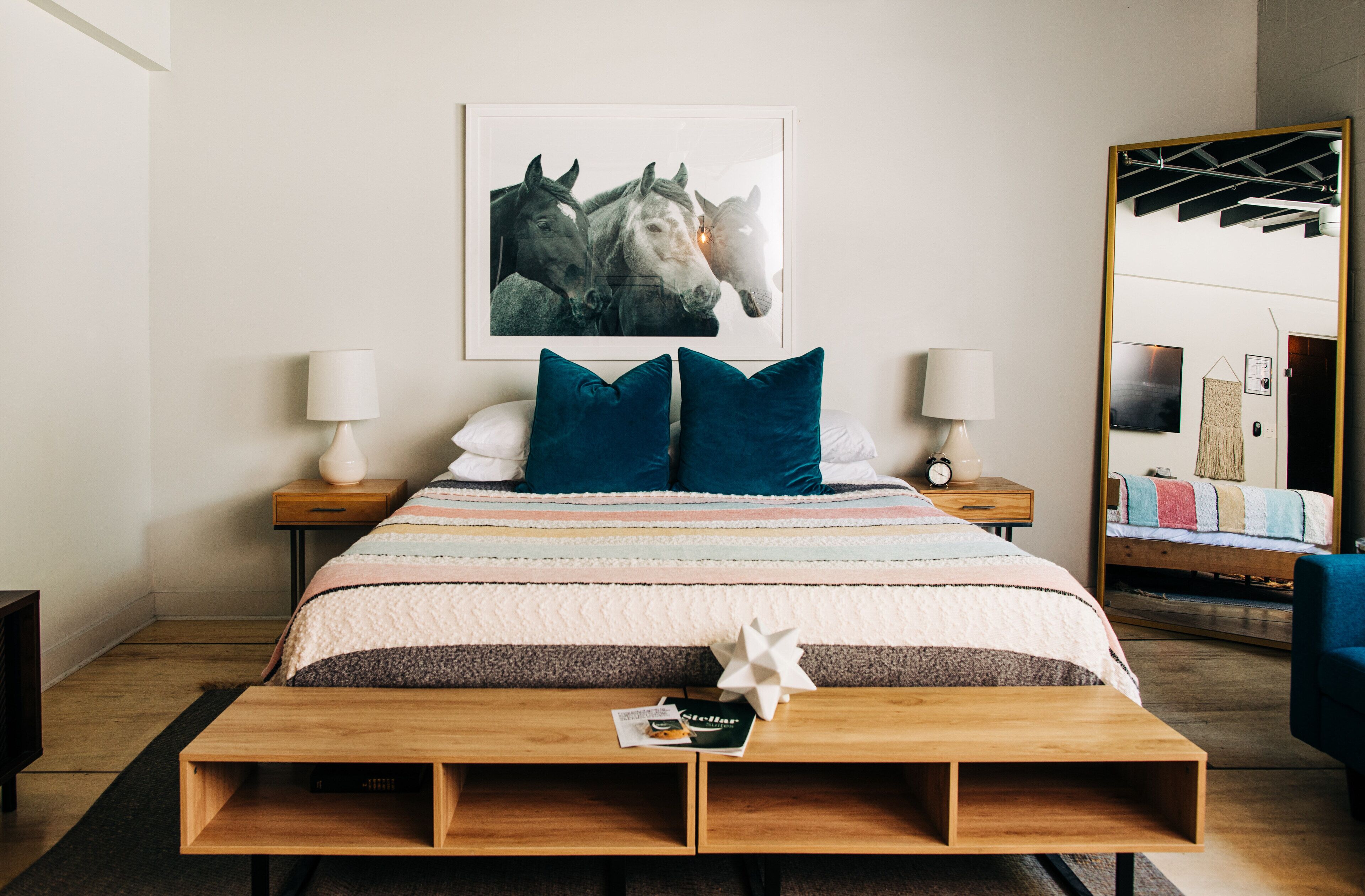 Une chambre à coucher bien rangée, avec un lit, des oreillers, des tables de chevet, une lampe, un miroir et un tableau encadré au mur.