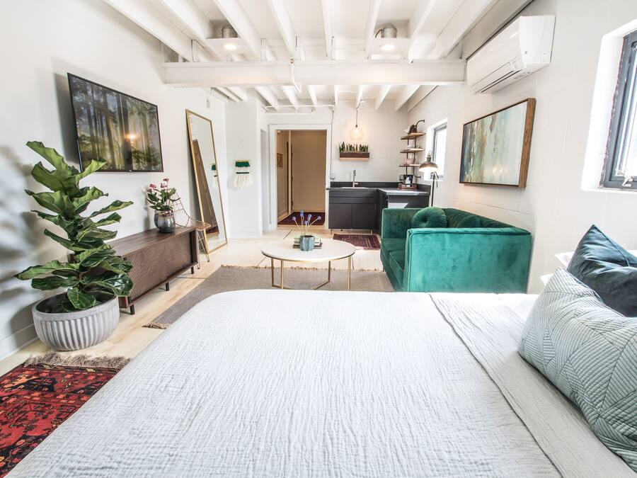 Una habitación moderna y bien iluminada con una cama grande, un sofá verde, una mesa redonda pequeña y una cocina al fondo.