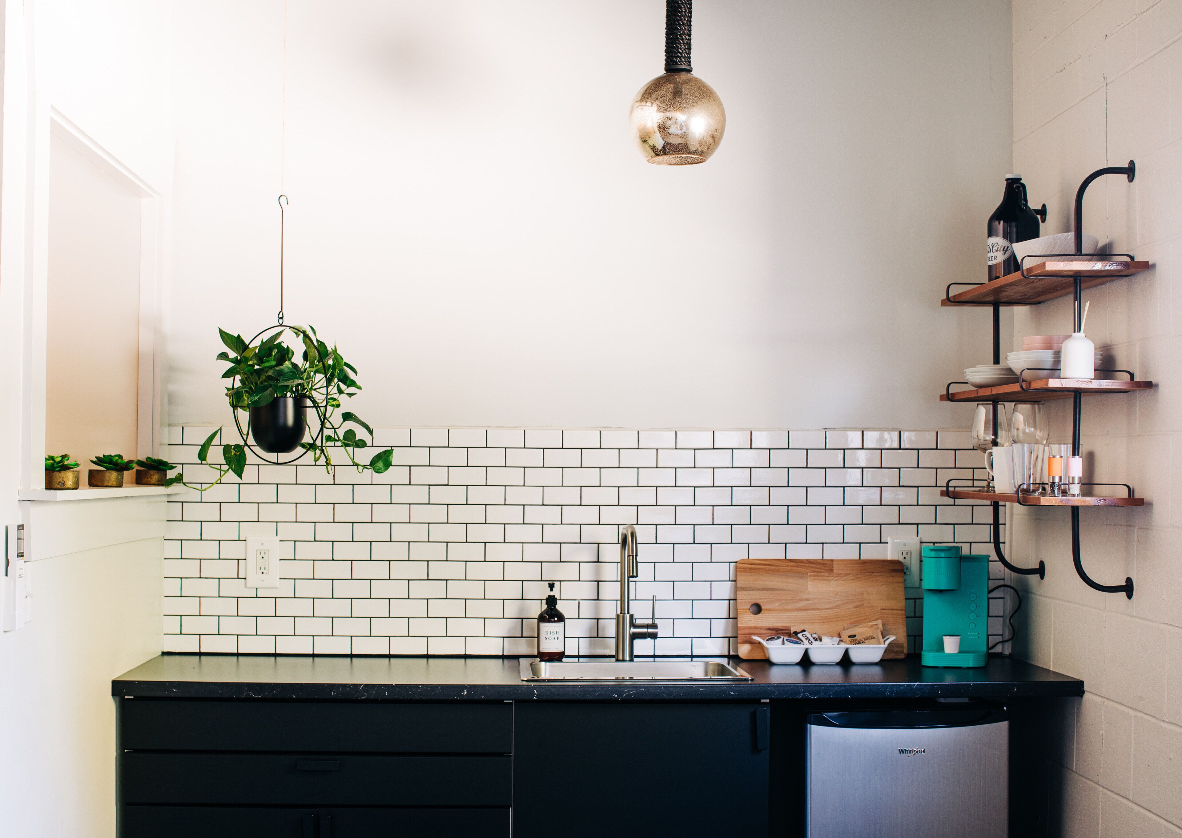 A kitchen with white subway tiles, a black countertop, a hanging plant, a wooden cutting board, and a coffee maker.