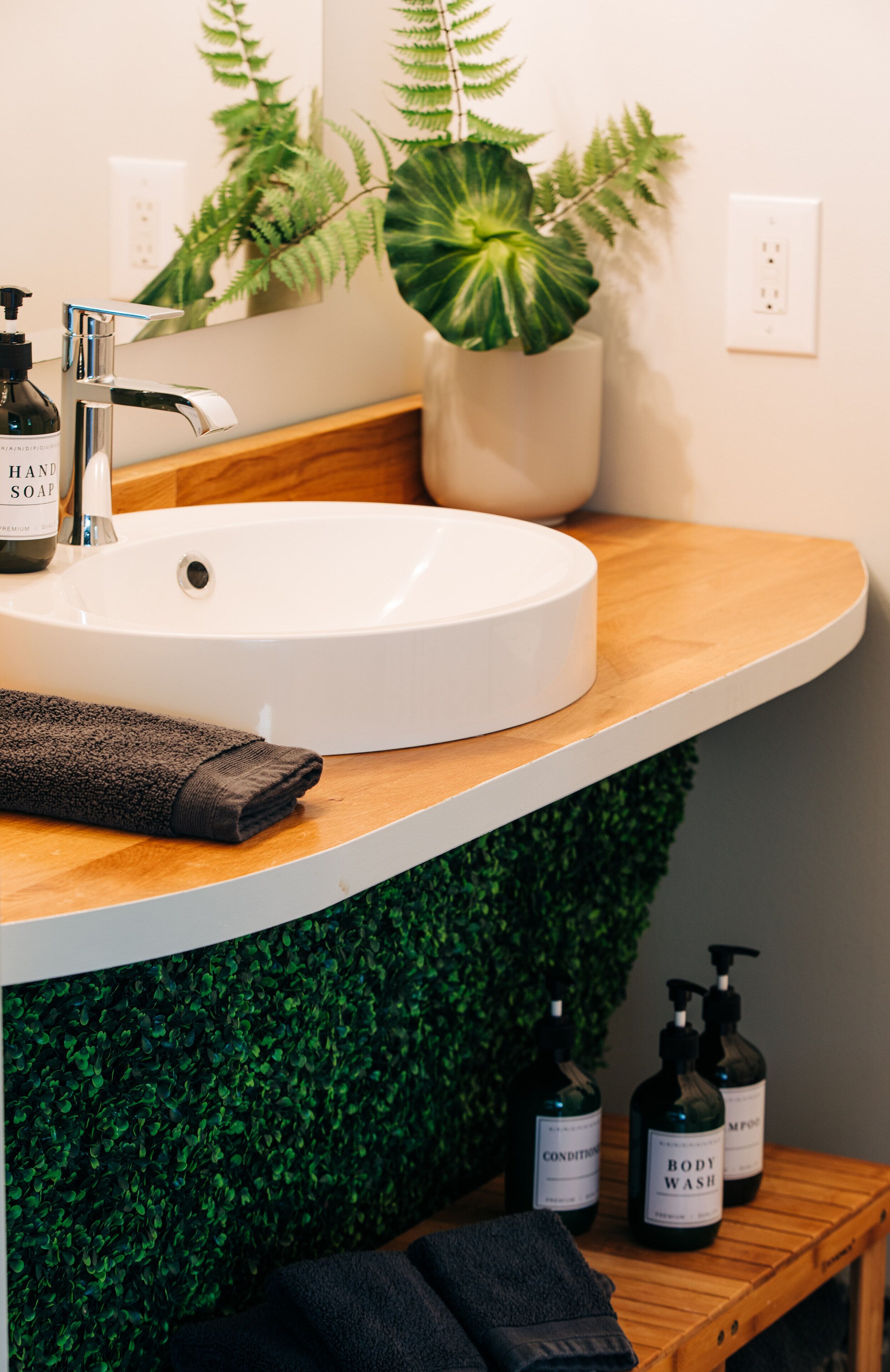 A bathroom with a white sink, wooden countertop, green foliage wall, and toiletries.