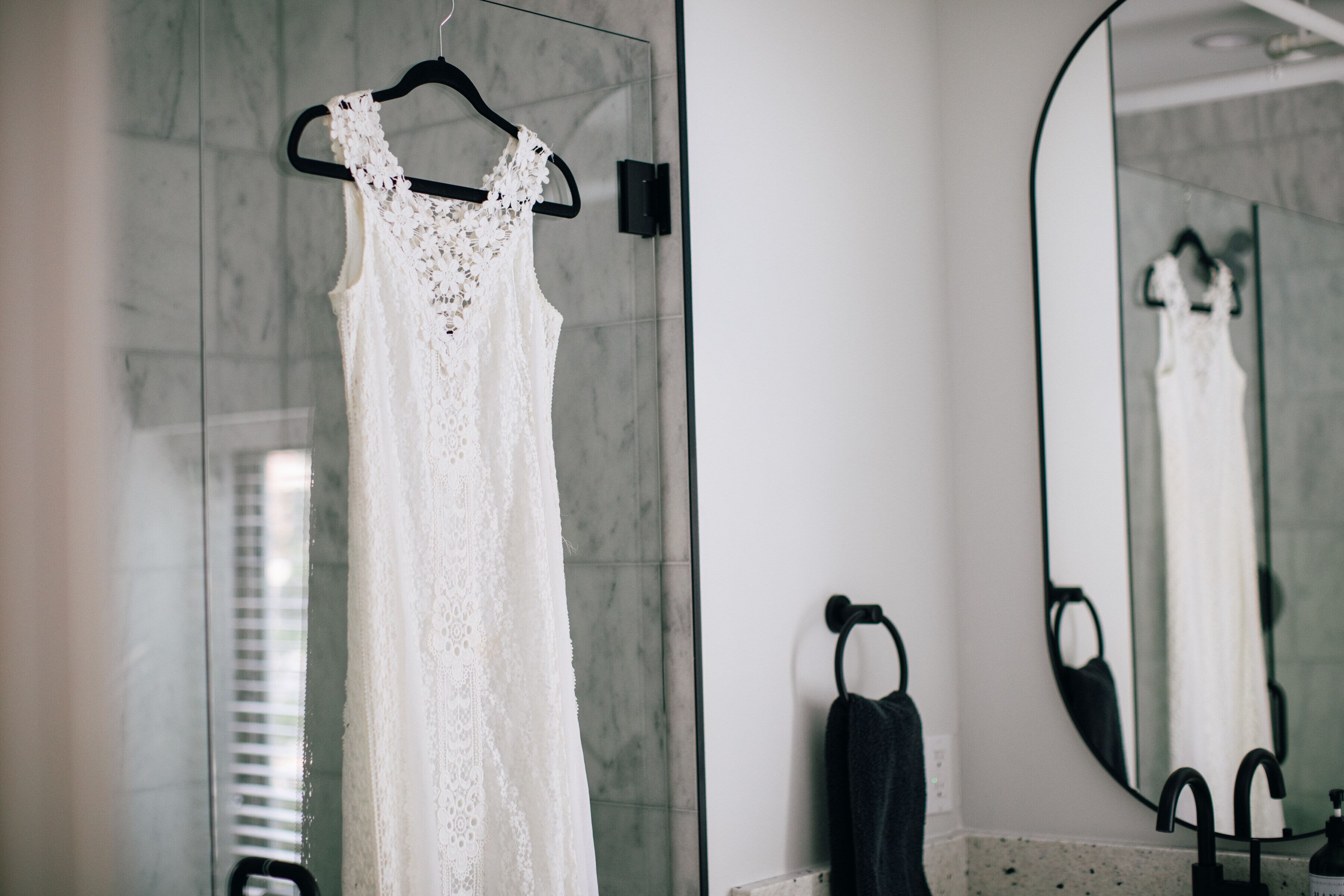A white lace wedding dress hanging on a black hanger in a bathroom with a large mirror, a black towel, and a marble countertop.
