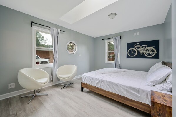 Ein Schlafzimmer mit einem Bett, zwei Stühlen, einem fensterbelaubten Fenster, einer Deckenleuchte und einem am Wand hängenden Fahrradplakat.