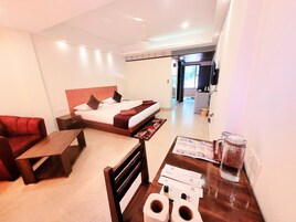 A hotel room with a bed, a desk, a chair, a sofa, and a table. There is a kitchen area visible through an open door.