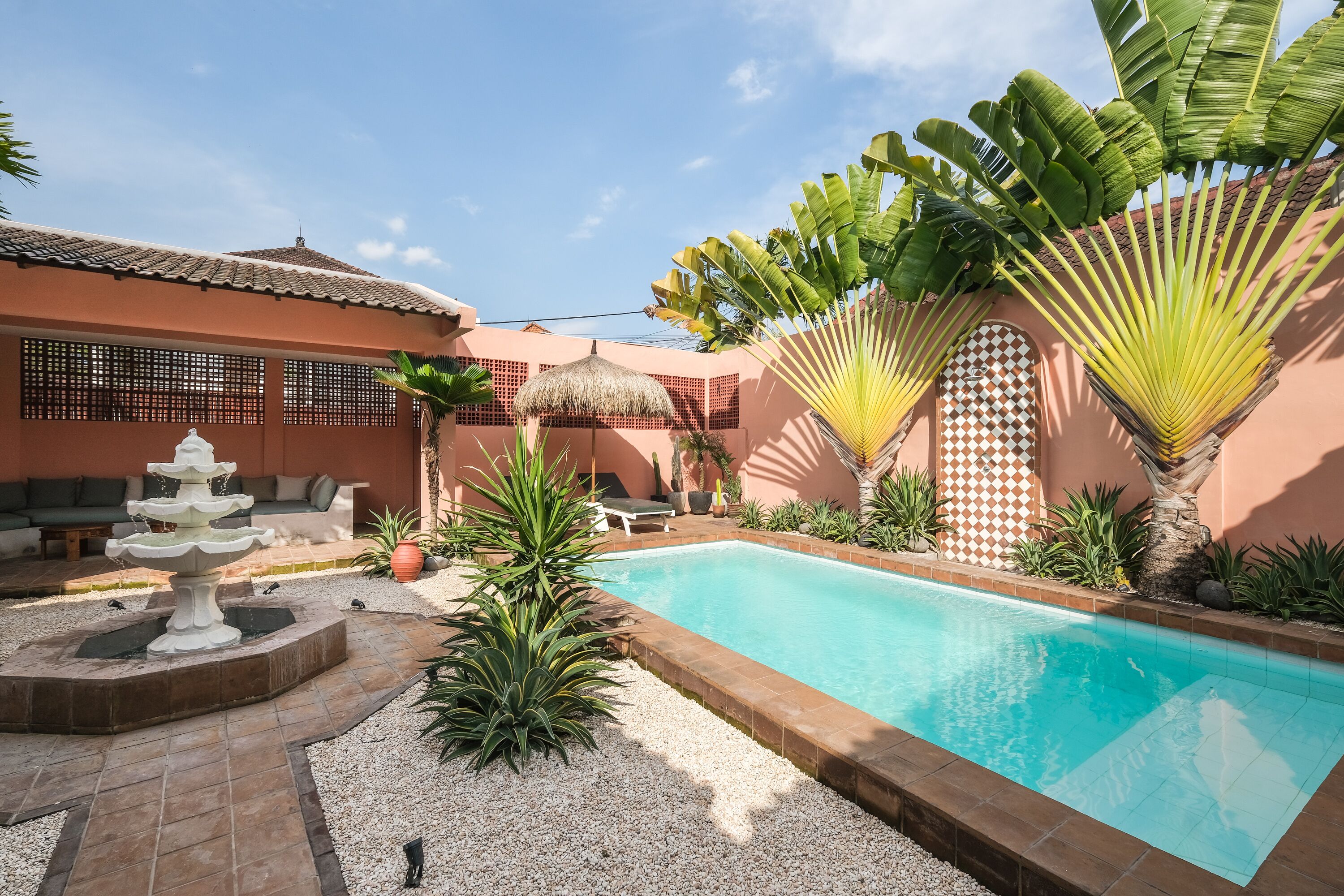 A swimming pool surrounded by a garden with a fountain, palm trees, and a thatched-roof structure.