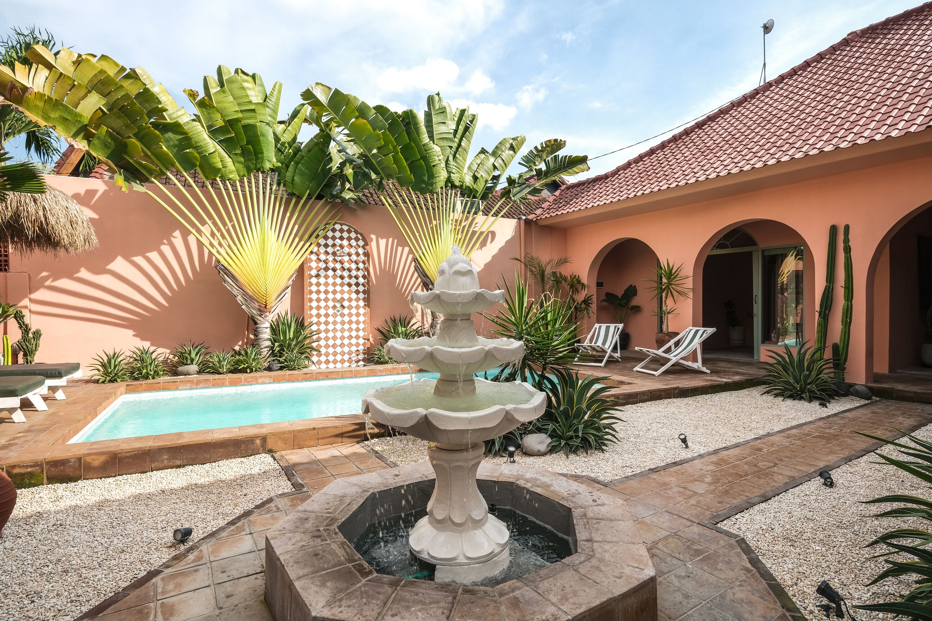 A pool area with a central fountain, surrounded by palm trees and a tiled patio.