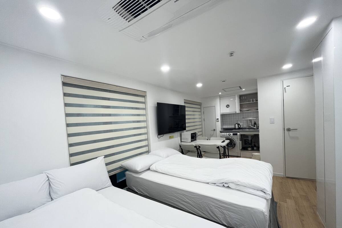 A modern hotel room with two beds, a small kitchenette, a flat-screen TV, and a large window with blinds.