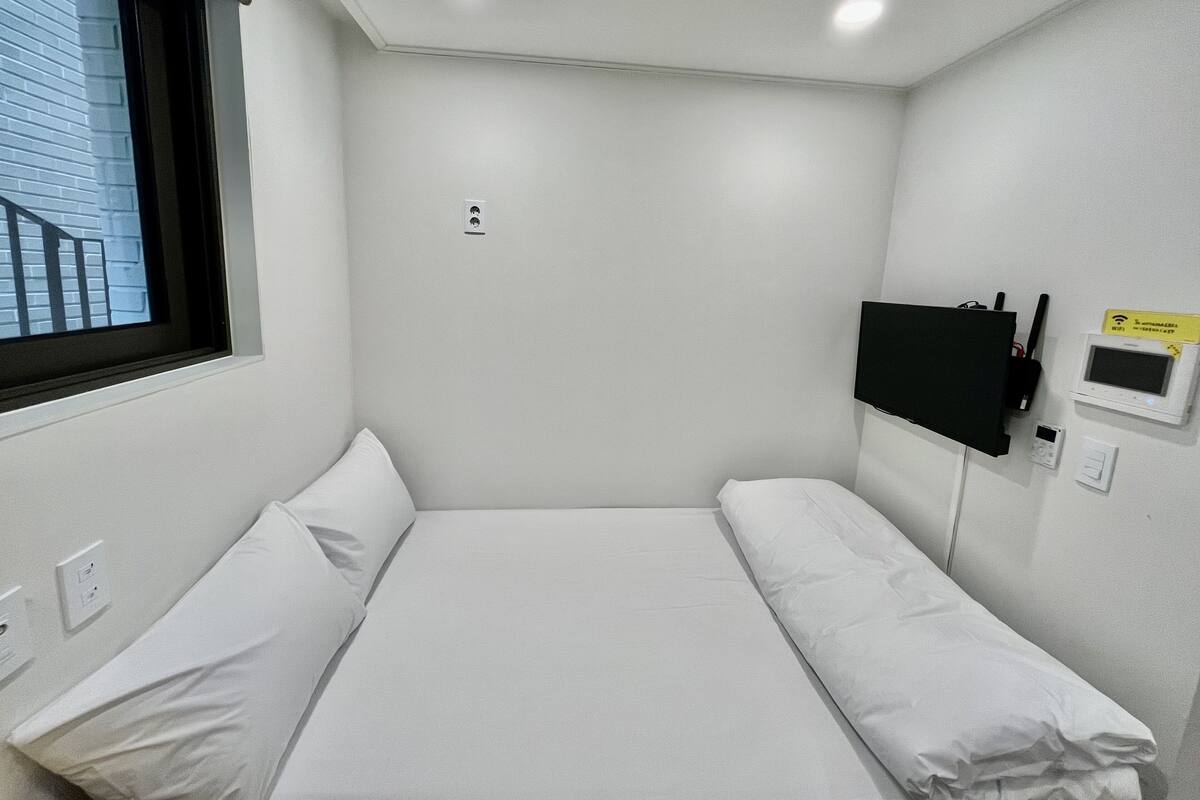 A small, single-bed room with a window, a flat-screen TV, a Wi-Fi router, and a power outlet.