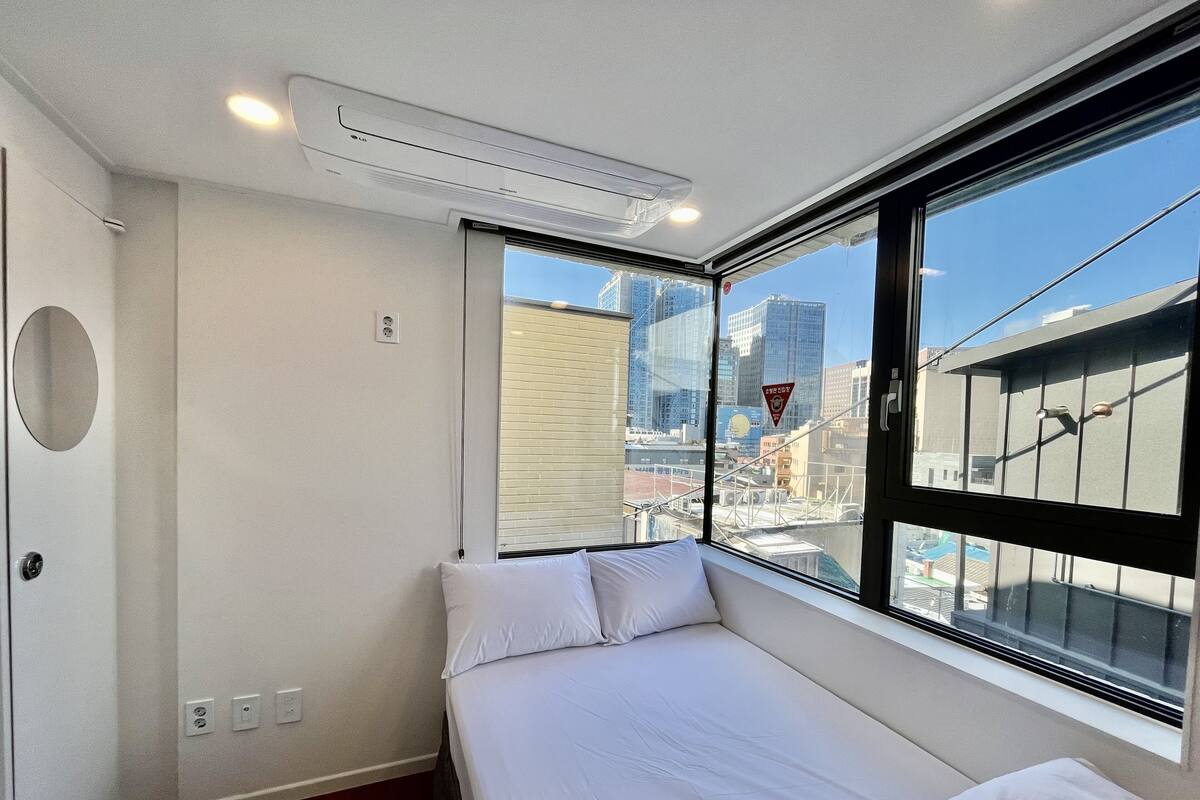 A hotel room with a bed, a window with a view of the city, and an air conditioning unit on the ceiling.
