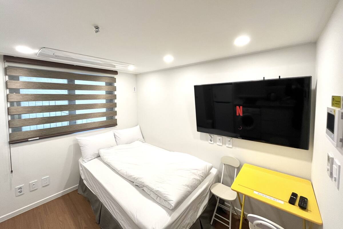 A hotel room with a bed, a TV mounted on the wall, a small desk with a chair, and a window with blinds.