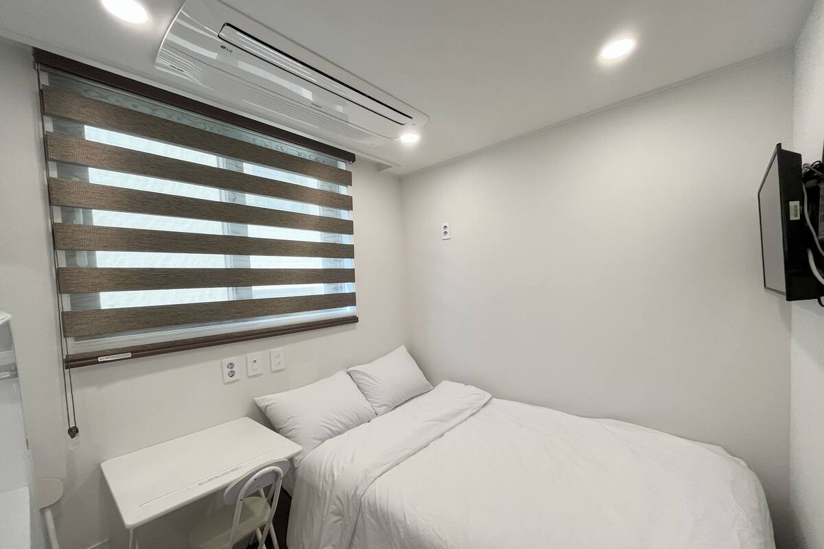 A bedroom with a bed, a desk, and a chair. There is a window with blinds, an air conditioning unit, and a wall-mounted TV.