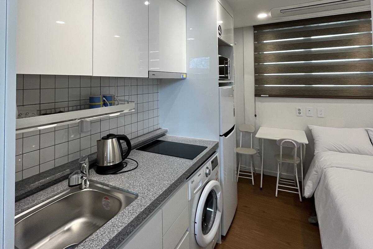 A compact kitchenette with a sink, stove, and washing machine, a small table, and a bed in a narrow room.