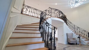 A grand staircase with ornate black railings, a beige armchair, and a chandelier. - Hilltop Mansion Near Saratoga @ Rebecca's Fountain with Chef, Masseur & Fun (Niskayuna)