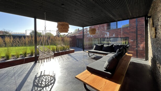Un espacio de estar moderno al aire libre con un banco de madera y cojines, una lámpara colgante, y vistas a un edificio de ladrillo y follaje.