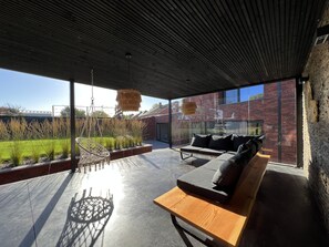 A modern outdoor seating area with a wooden bench and cushions, a hanging light fixture, and a view of a brick building and greenery. - Group home with swimming pool and wellness (Heers)