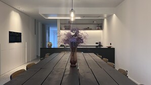 Un comedor moderno con una larga mesa de madera, un florero con flores moradas y una cocina al fondo.
