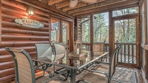 A wooden cabin interior with a welcome sign, a glass table, metal chairs, and large windows offering a view of the forest. - Aska Adventure Cabin on 3 Acres • Hot Tub • Screened Decks • Game Loft (Cherry Log)