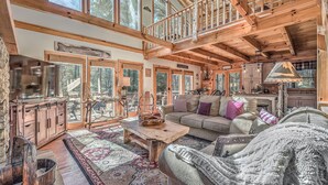 A spacious cabin interior with a high ceiling, wooden beams, a large window overlooking a forest, a cozy seating area with a sofa and armchairs, and a rustic kitchen with wooden cabinets. - Aska Adventure Cabin on 3 Acres • Hot Tub • Screened Decks • Game Loft (Cherry Log)