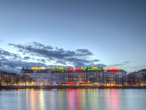 A waterfront hotel with multiple stories, a river in the foreground, and illuminated signage.