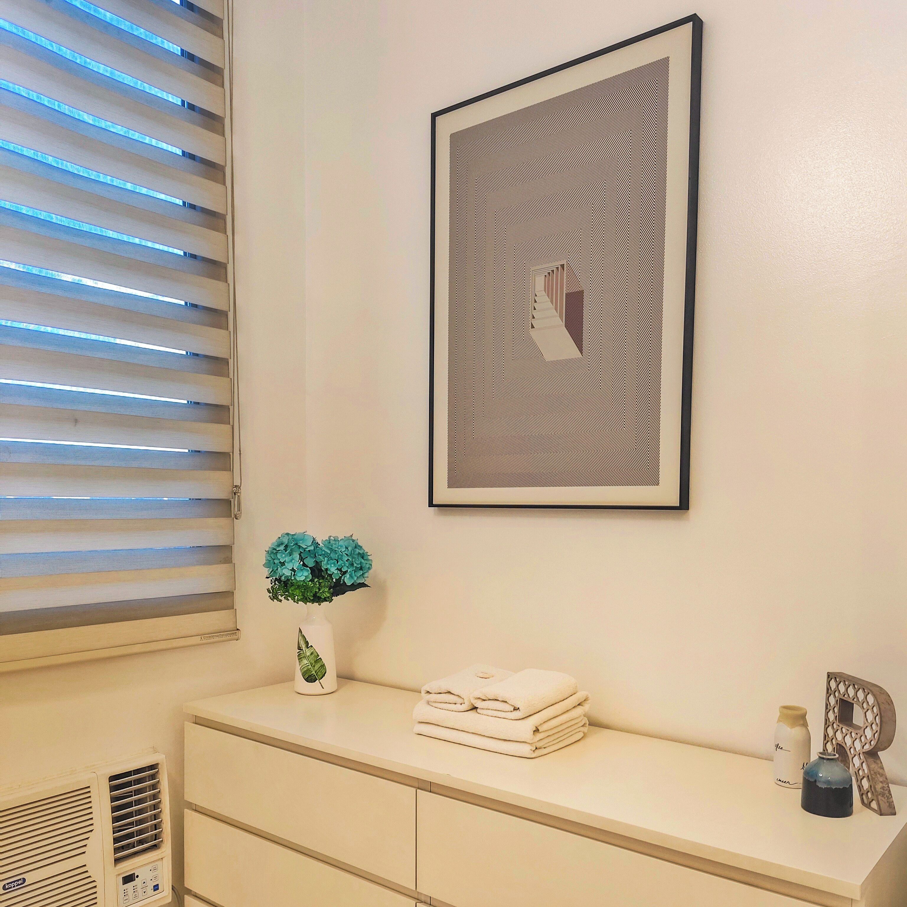A bathroom with a vanity, a framed artwork, a vase with flowers, and a towel stack.