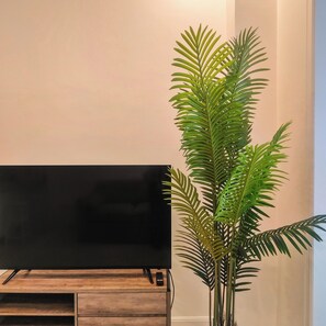 A flat-screen TV on a wooden stand, a large potted plant, and a white wall. - Voss Condominium (Quezon City)