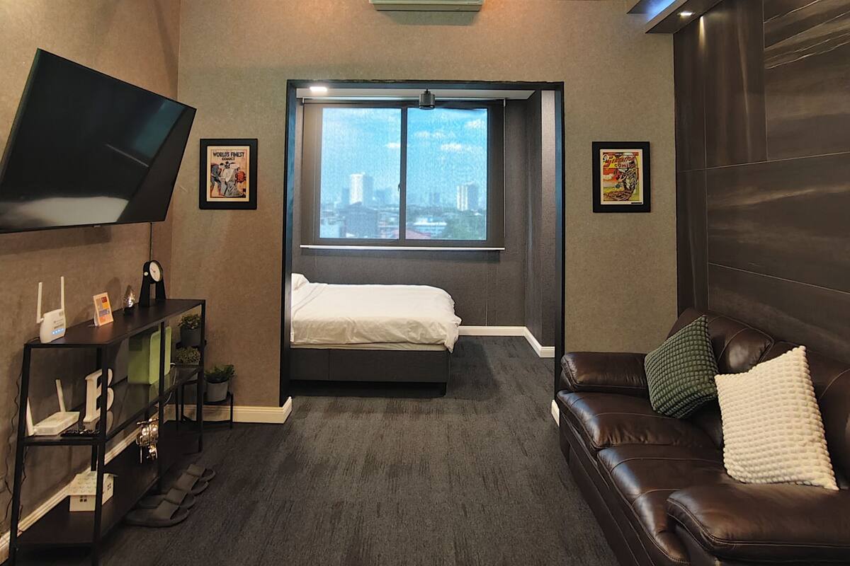 A modern hotel room with a bed, a TV, a window with a city view, a sofa, and a small table with a router and decorative items.
