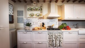A kitchen with white cabinets, a tiled backsplash, and a stove. There are plates on a shelf above the stove and a plant on the counter. - Podere Il Chiesino 8 2 (Montecatini Val di Cecina)