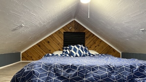 Ein Schlafzimmer mit schräg verlaufender Decke, Holzvertäfelung, einem Bett mit geometrischem Muster auf der Bettdecke und einer einzelnen Pendelleuchte.