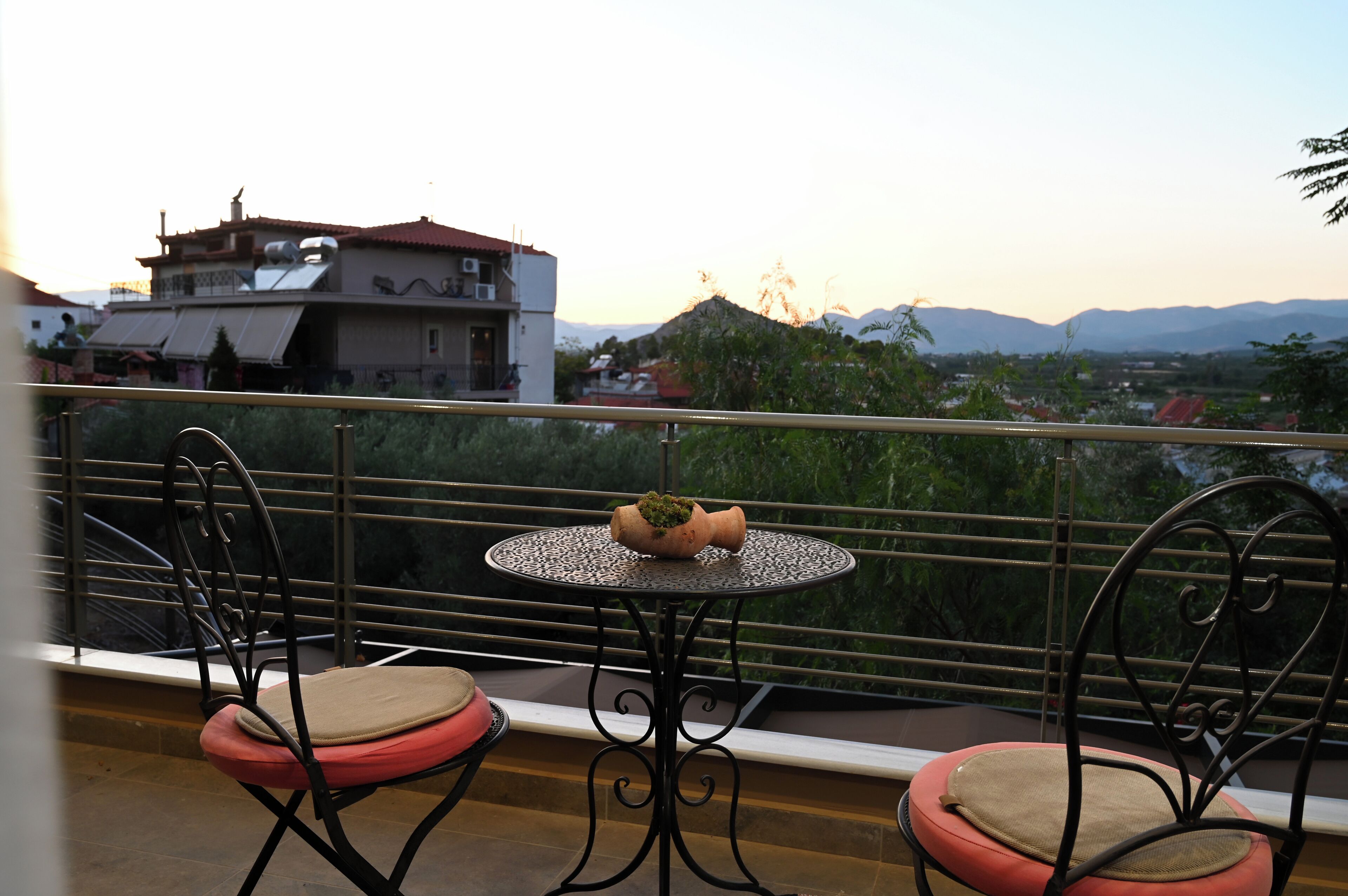 Un balcon avec une table ronde et deux chaises, un pot de fleurs et une vue sur des montagnes et des bâtiments éloignés.