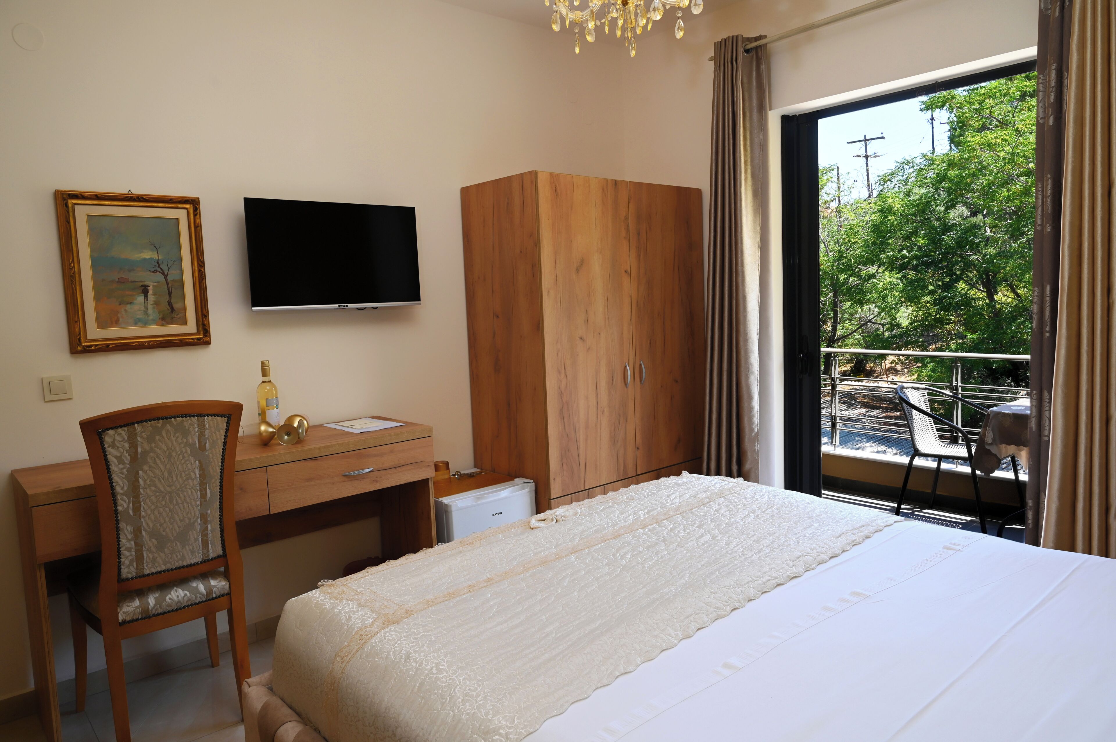 A hotel room with a bed, a desk with a chair, a television, a wardrobe, and a balcony with a view of trees.