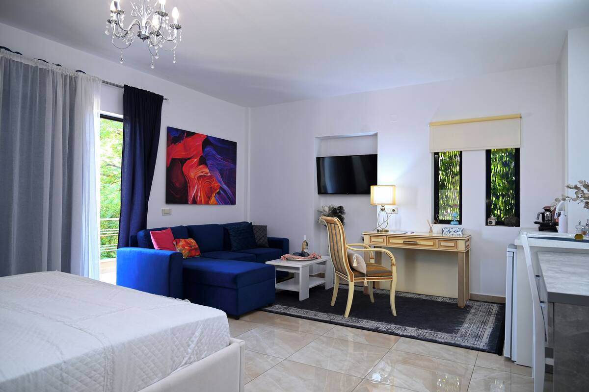 A hotel room with a bed, a blue sofa, a desk with a chair, a TV, and a window with curtains.