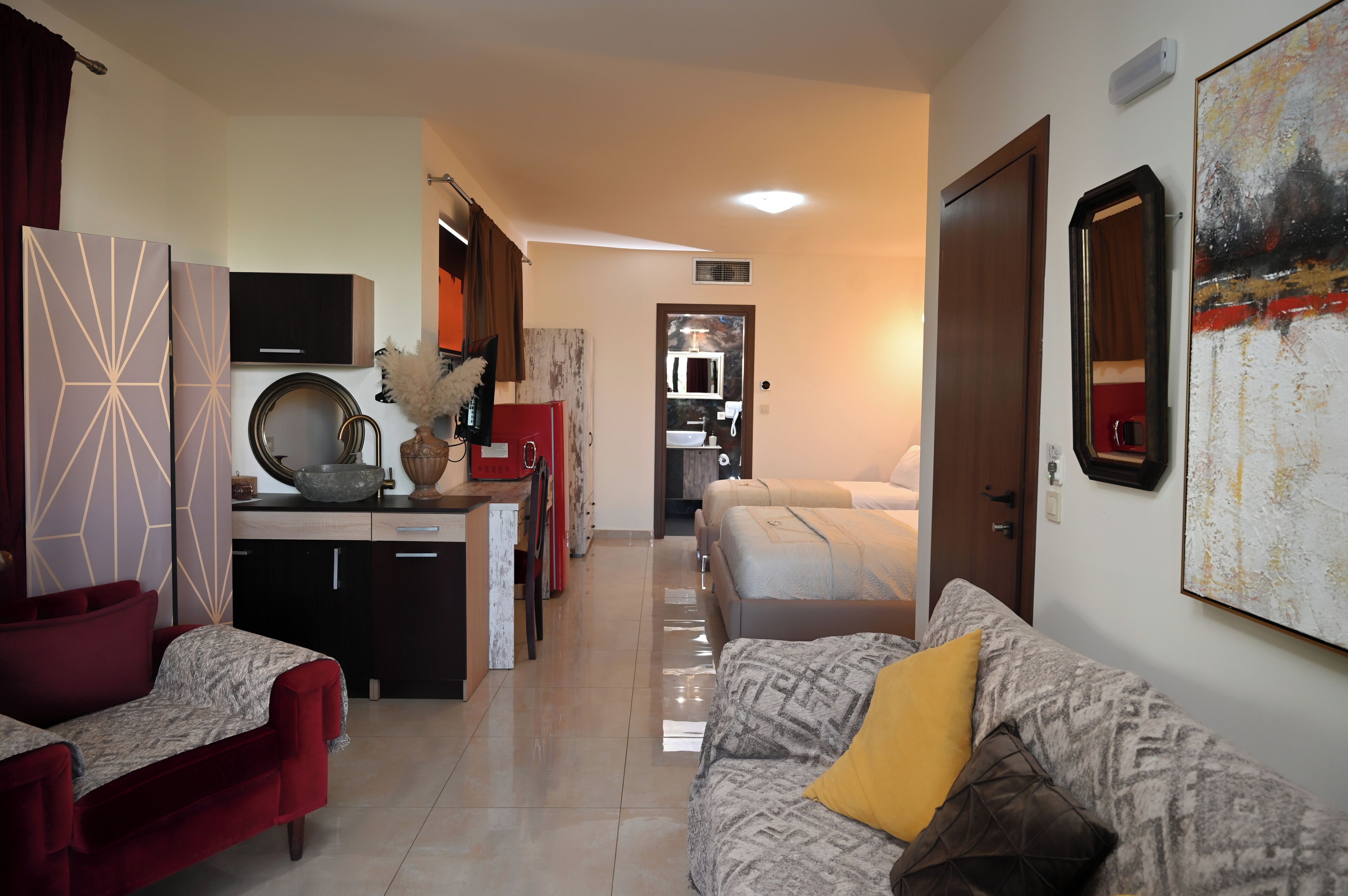 A modern hotel room with a kitchenette, a dining area, and a living space with a sofa and a red chair.