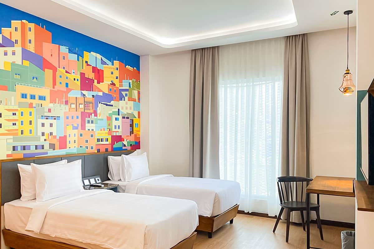 Ein Hotelzimmer mit zwei Betten, einem Schreibtisch, einem Stuhl, einem großen farbenfrohen Wandgemälde und einem Fenster mit Vorhängen.