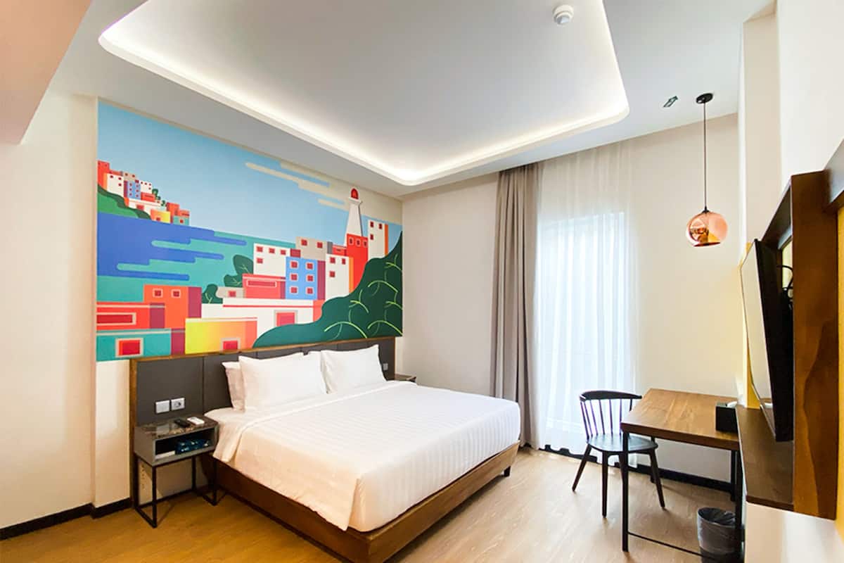 Ein modernes Hotelzimmer mit einem großen Bett, einem Schreibtisch und einem Sessel. An der Wand hängt ein Gemälde, das eine farbenfrohe Stadtansicht zeigt.