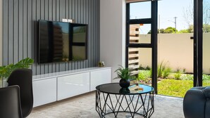 Un salon moderne avec un téléviseur à écran plat fixé au mur, un canapé noir, une table basse ronde et une grande fenêtre donnant sur l’extérieur.