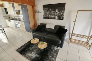 A modern living room with a black leather sofa, two round wooden side tables with plants, a black shag rug, a cityscape painting, and a wooden rack.
