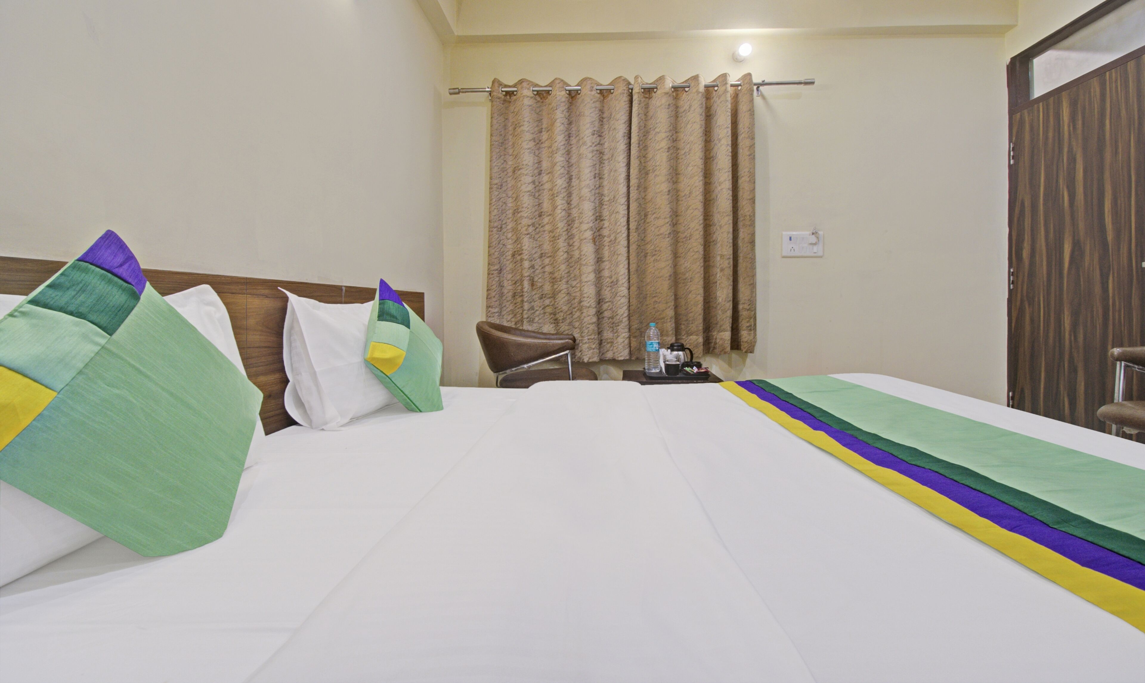 A hotel room with a bed, pillows, a chair, a nightstand, and a window with curtains.