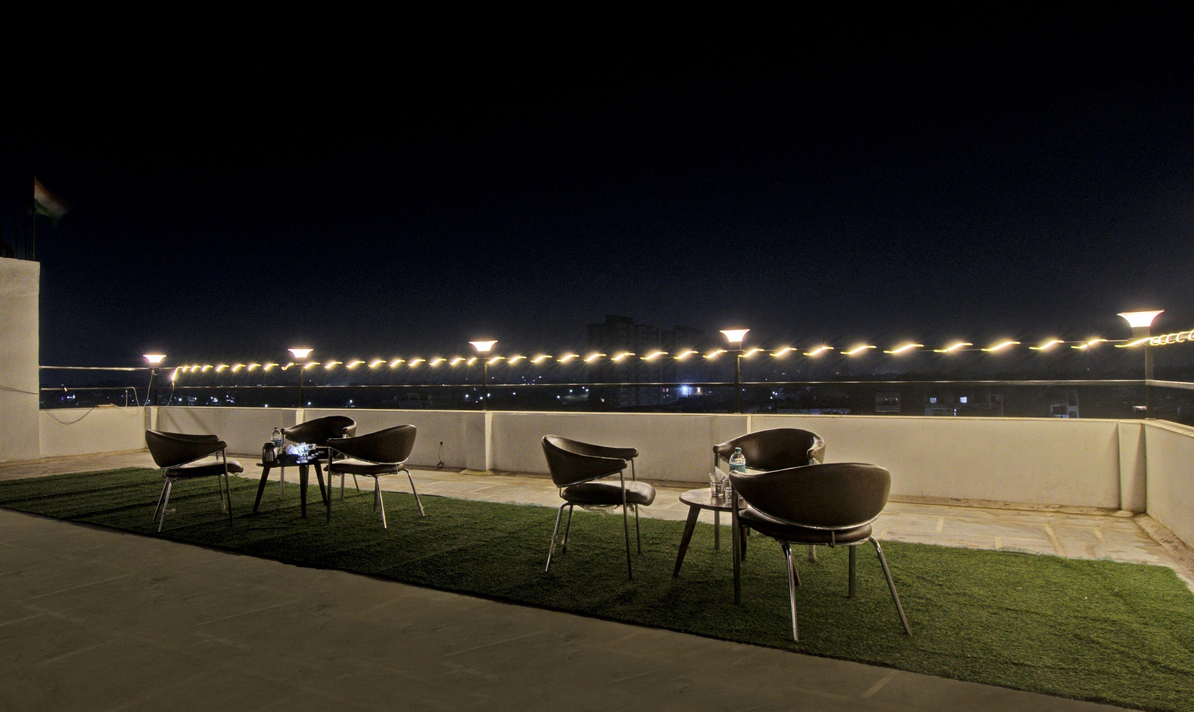 A rooftop terrace with chairs and tables, string lights, and a view of the cityscape at night.