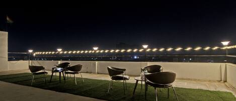 A rooftop terrace with chairs and tables, string lights, and a view of the cityscape at night.