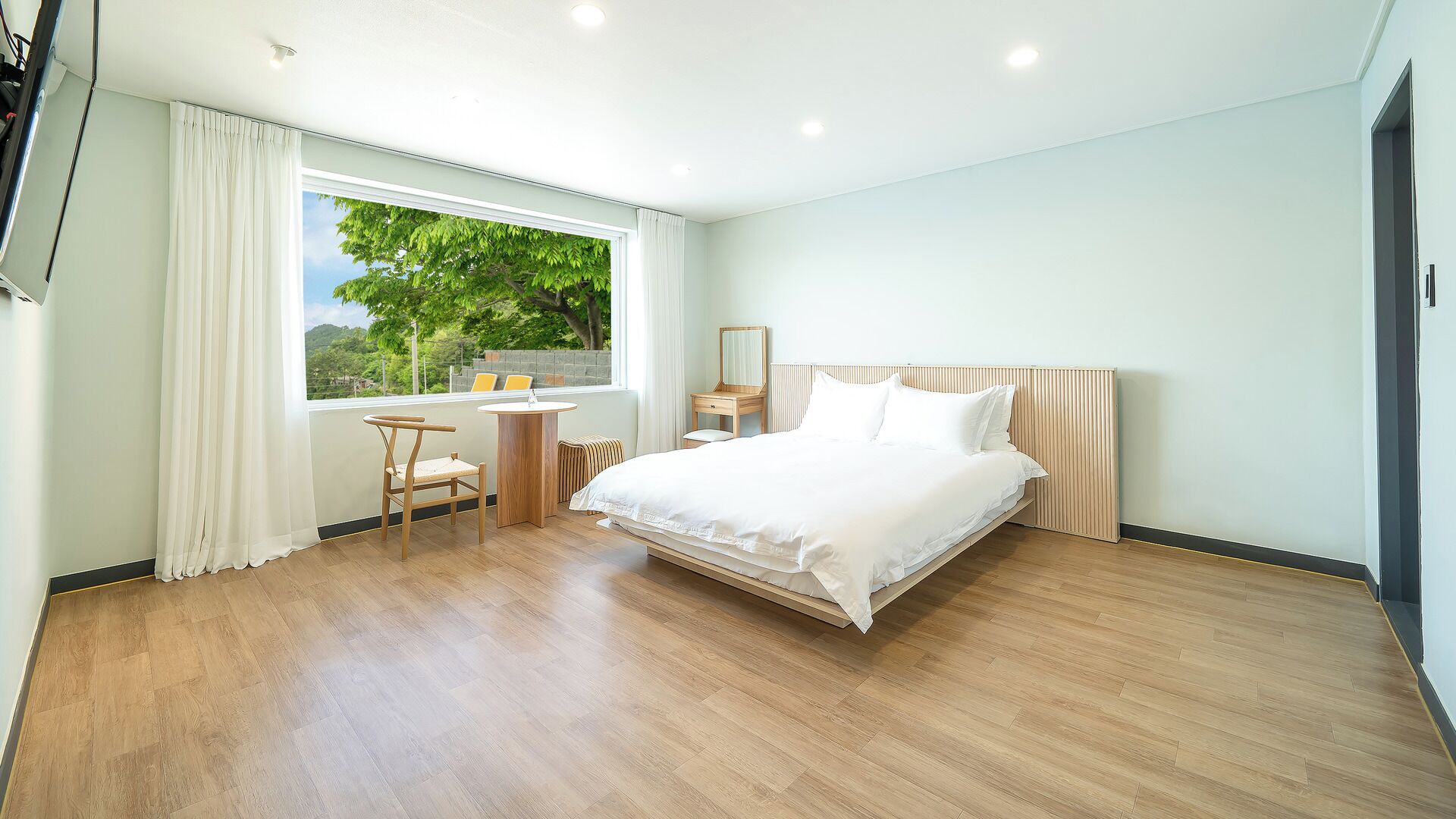 A modern hotel room with a large bed, a small desk, a chair, and a window with a view of trees.
