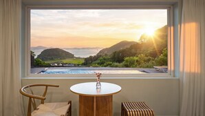 A room with a view of a coastal landscape, a round wooden table, a chair, and a small stool.